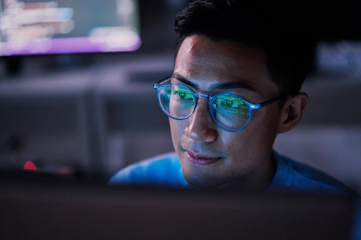 Developer wearing blue glasses viewing code on computer screen in dark office environment